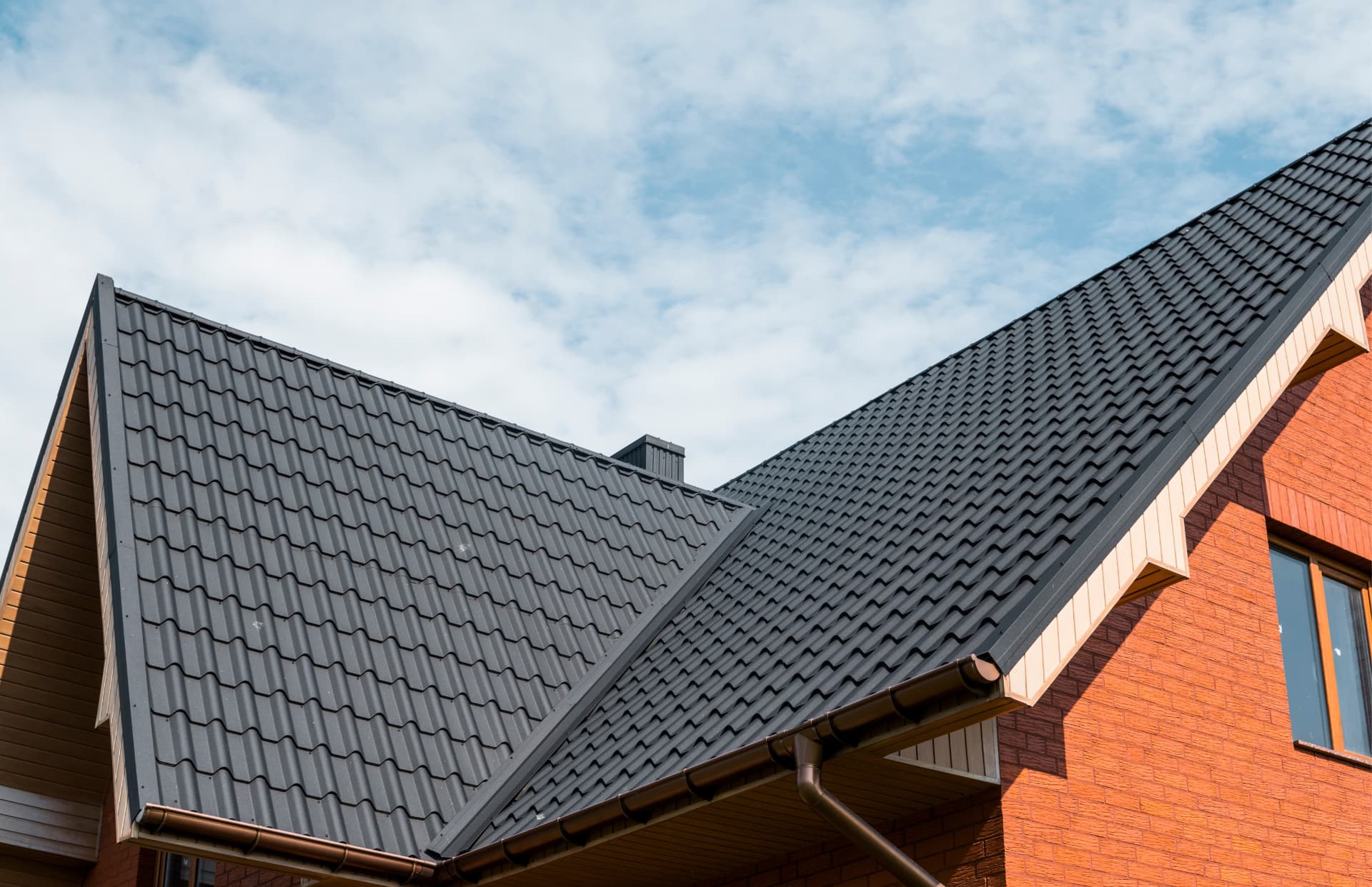 Close-up of a dark metal shingle roof on a brick house beneath a partly cloudy sky
